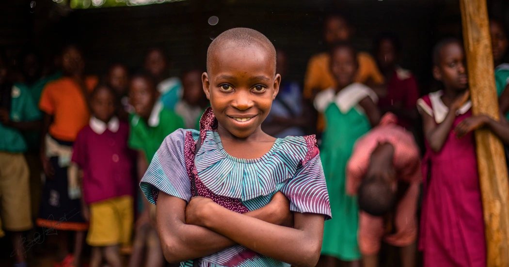Confident Ugandan schoolgirl stands with folded arms, gazing into the camera as classmates look on—symbolizing the hope and strength Baino Social Impact nurtures through education in underserved communities.