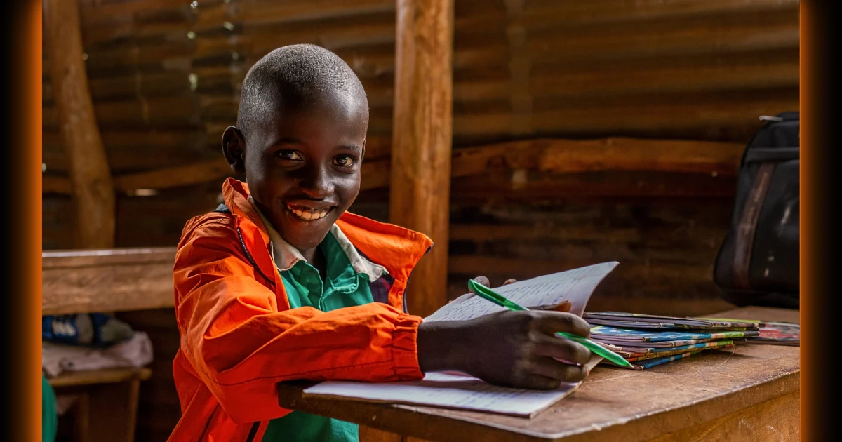 A smiling Ugandan boy proudly holds up his pen in a worn rural classroom, capturing the hope and determination Baino Social Impact fights to build and protect through education and child empowerment.