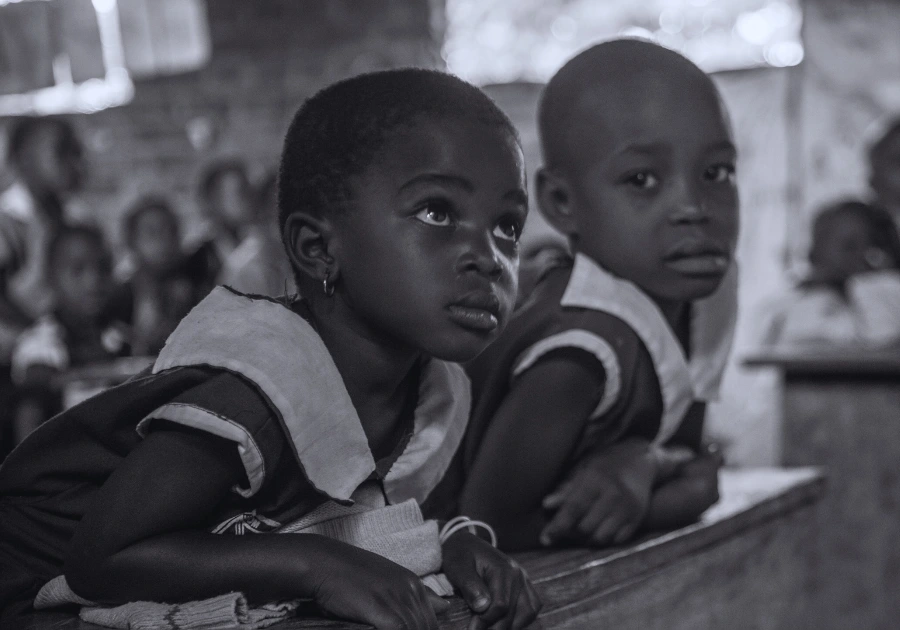 Two young Ugandan kindergarten girls in a rural classroom—one watching the teacher, the other looking into the camera—capturing the innocence, curiosity, and promise that Baino Social Impact strives to protect through transparent, accountable education programs.