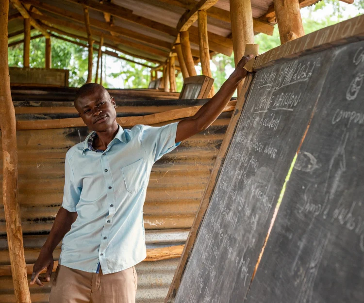 A dedicated teacher stands by a movable blackboard in a rural Ugandan classroom made of iron sheets, guiding students with care and commitment to learning.