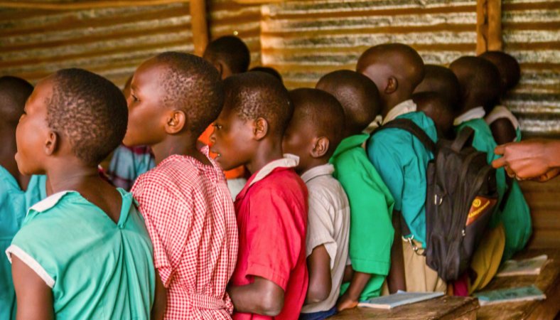 Side view of children seated at a long desk in a crowded rural classroom, facing the blackboard and singing, illustrating eagerness to learn and the challenges of underserved education addressed by Baino Social Impact.