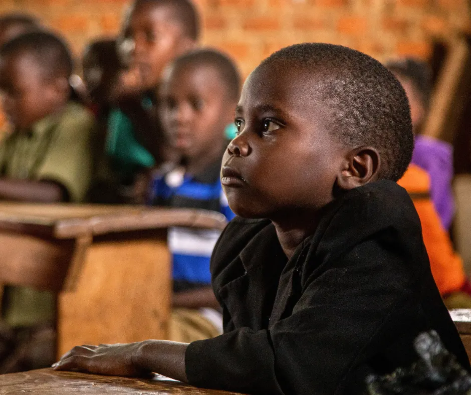 Young Ugandan girl listens intently to her teacher in a modest classroom, surrounded by blurred classmates—capturing the hope and determination Baino Social Impact nurtures through access to quality education.
