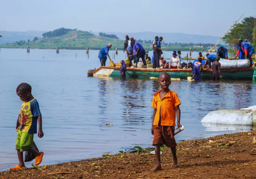 Two rural Ugandan boys walk barefoot and in slippers along the shore of Lake Victoria, while workers unload items from a boat in the background. The scene reflects the challenges children face when schooling is out of reach, emphasizing the urgent need for educational support through Baino Social Impact.
