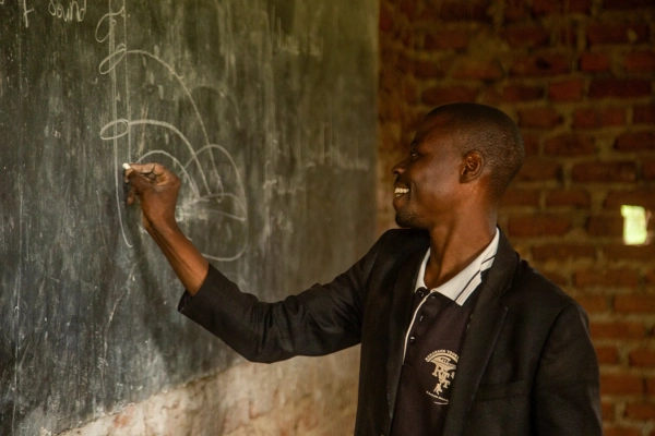 A rural Ugandan music teacher writes a musical note on a blackboard in an unfinished classroom with exposed bricks. The image highlights the dedication of teachers and the need for educational support, showing how Baino Social Impact turns scarce resources into opportunities for learning and hope.