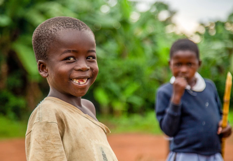 A smiling rural boy looks at the camera while a girl enjoys sugarcane in the blurred background, capturing the joy and resilience of children whose futures we can transform together—stand with us to support education in Uganda’s poorest communities.