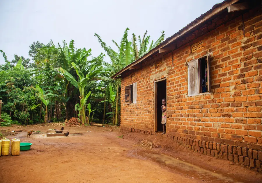 Shy Ugandan girl peeps from the doorway of her rural home as chickens feed in the compound—a quiet glimpse into the everyday realities Baino Social Impact seeks to transform through education and opportunity.