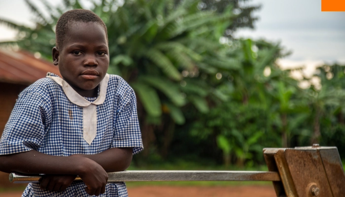 A rural Ugandan schoolgirl in a worn blue-and-white uniform holds a borehole pump, reflecting the daily challenges faced by children in underserved communities and Baino Social Impact’s mission to bring education and dignity closer to home.