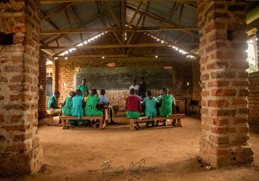 Long shot of a rural Ugandan classroom with an unfinished brick building, dirt floor, and a few students listening to their teacher—reflecting the humble realities Baino Social Impact works to transform through transparency, education, and hope.