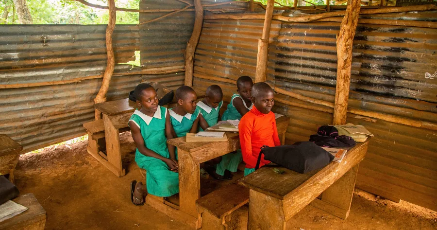 A group of young Ugandan schoolgirls sit attentively in a rural classroom with makeshift iron-sheet walls and bare-dirt floors, symbolizing the urgent need for better education resources in Uganda’s poorest communities.