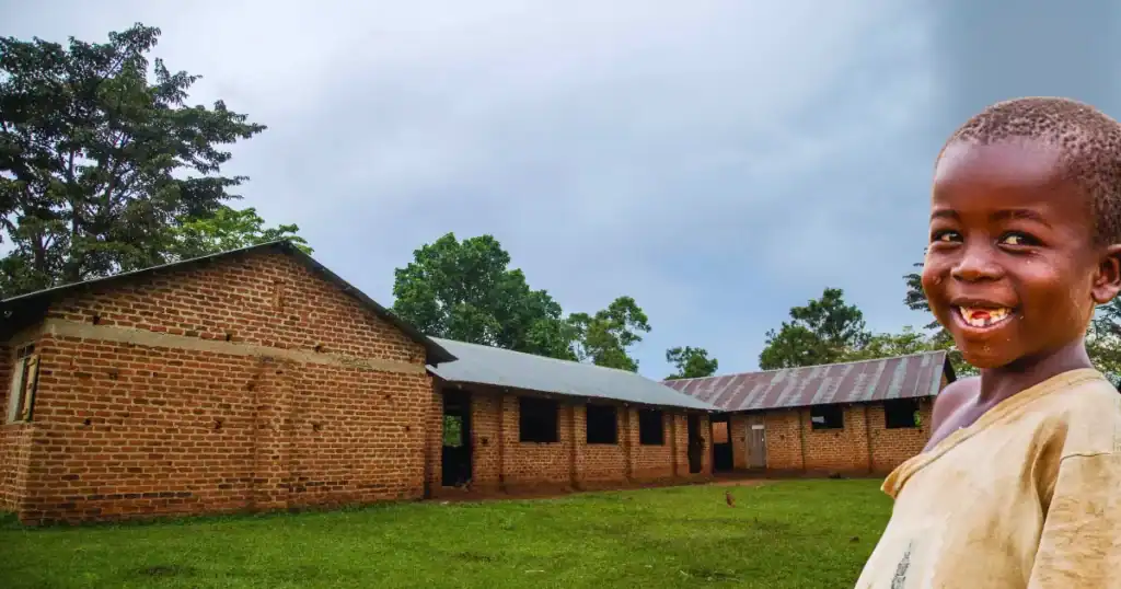 A smiling young boy stands before an unfinished school in rural Uganda — a living symbol of hope, progress, and the promise that every brick laid brings education closer to home.