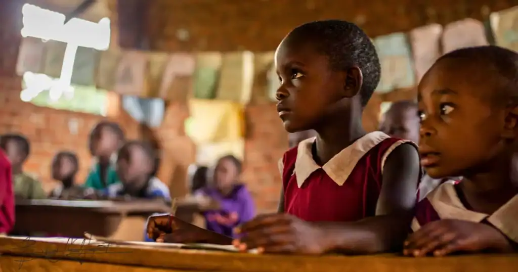Close-up of two young girls attentively listening in a rural Ugandan classroom — a tender reminder of why Baino Social Impact invites supporters to help lay the next brick and keep building safe, hopeful spaces where children can learn and thrive.