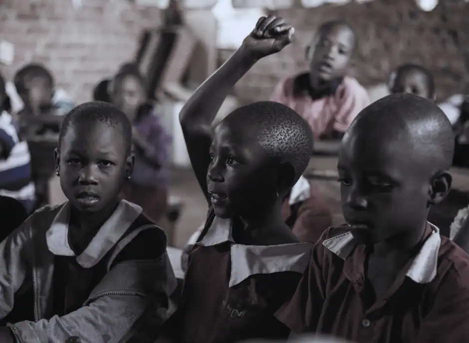 Young pupils learning in a rural Ugandan classroom, with one girl eagerly raising her hand to answer a question — capturing the spirit of confidence, curiosity, and renewed hope inspired by Baino Social Impact’s education projects.
